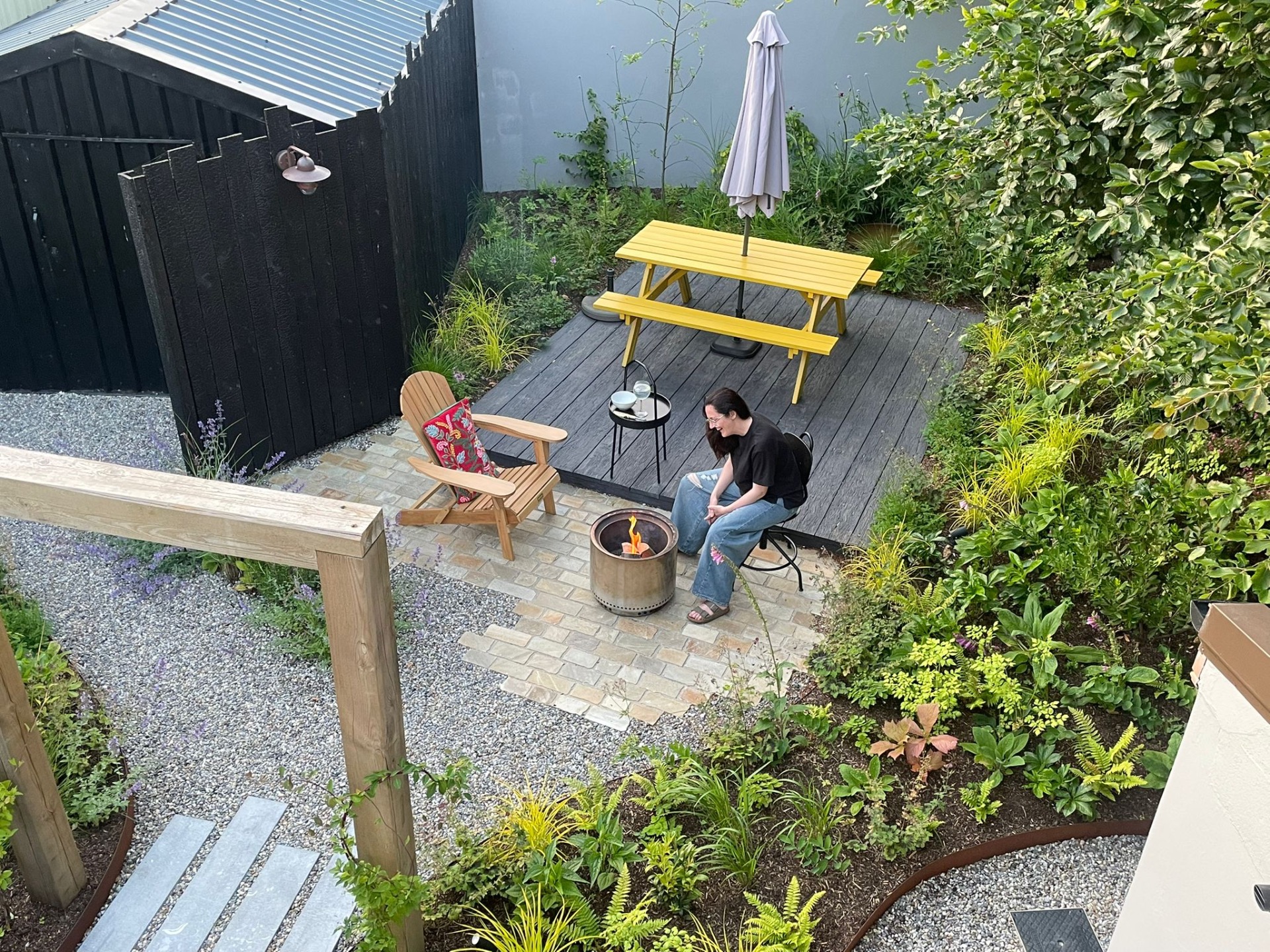 Woman sits on patio near fire pit, yellow table, and umbrella, surrounded by lush garden greenery.